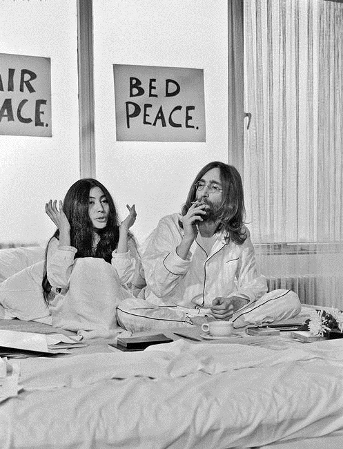 black and white photograph of two people sitting on a bed in front of a sign that says bed peace