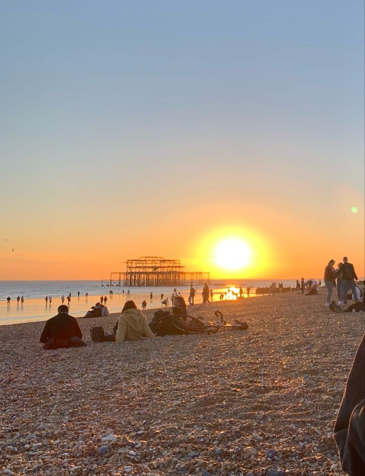 people are sitting on the beach watching the sun rise over the ocean and pier in the distance