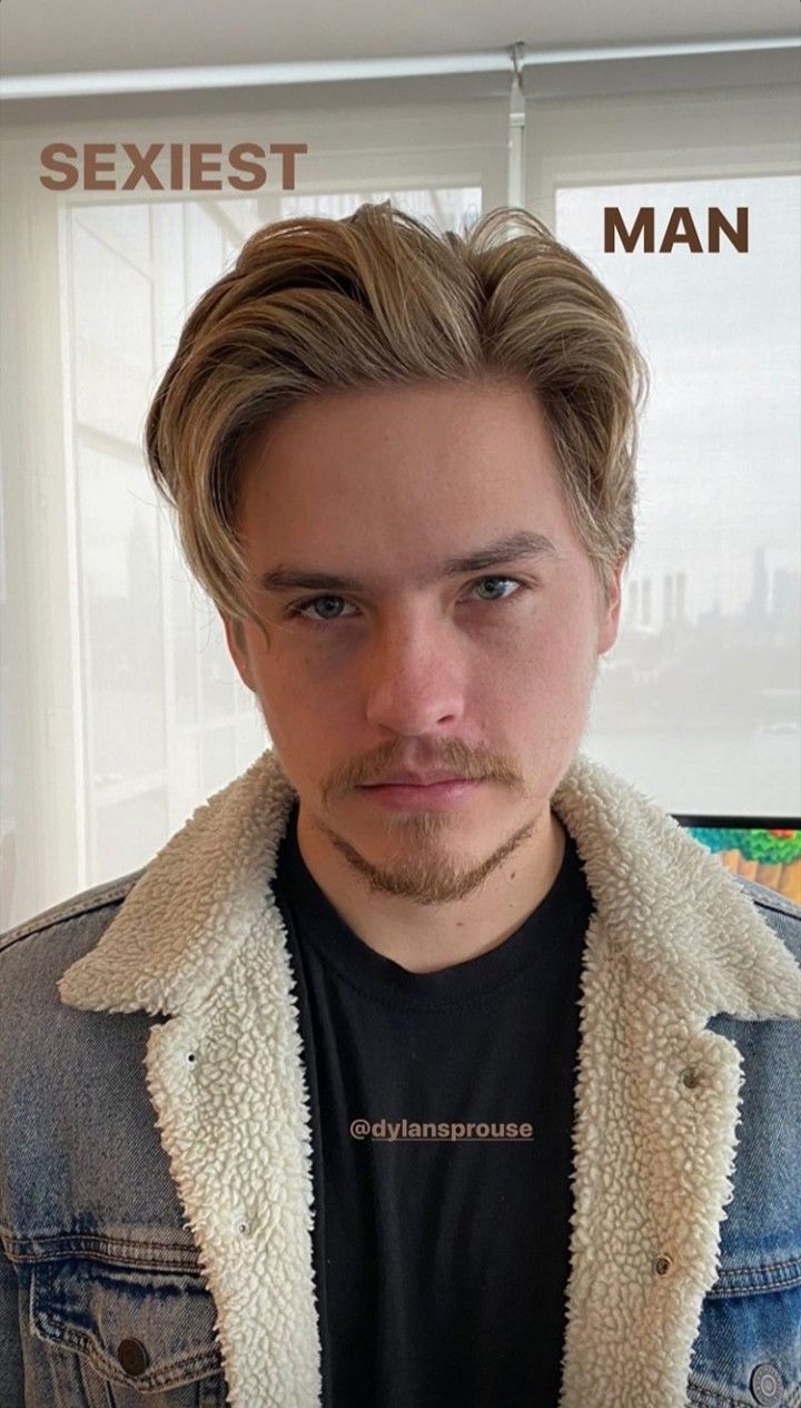 a man with long blonde hair wearing a denim jacket and looking at the camera while standing in front of a window