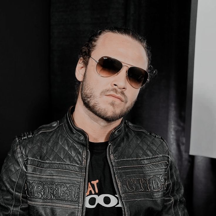 a man wearing sunglasses and a leather jacket sitting in front of a black curtain with his hands on his hips