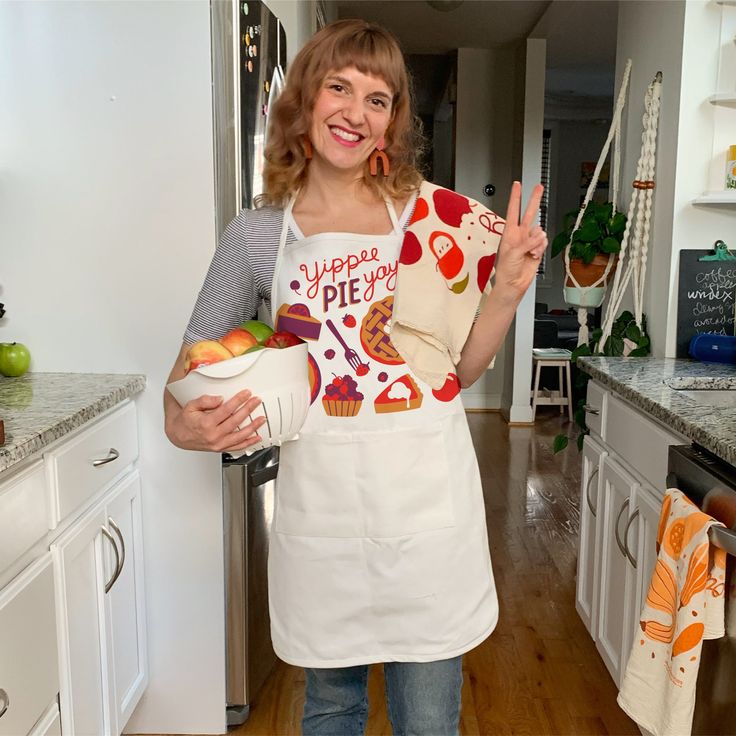 a woman in an apron is holding up her peace sign and some fruit on the counter