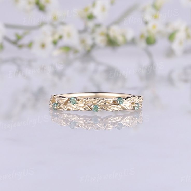 a gold ring with leaves and green stones on the side, sitting next to white flowers