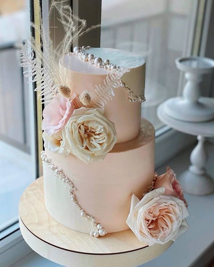 a three tiered wedding cake with flowers and feathers on the top, sitting in front of a window