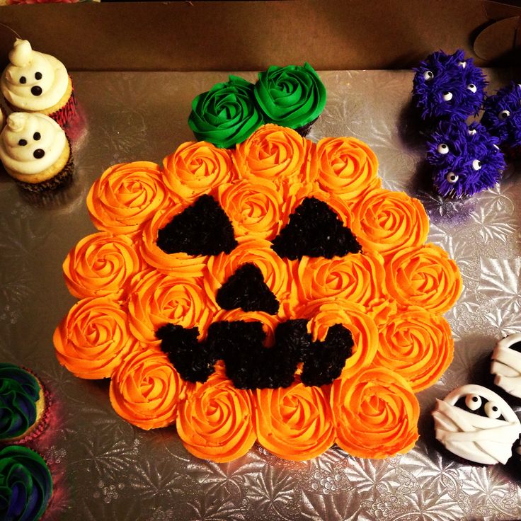 cupcakes with icing and decorations are arranged on a silver surface, including a pumpkin face