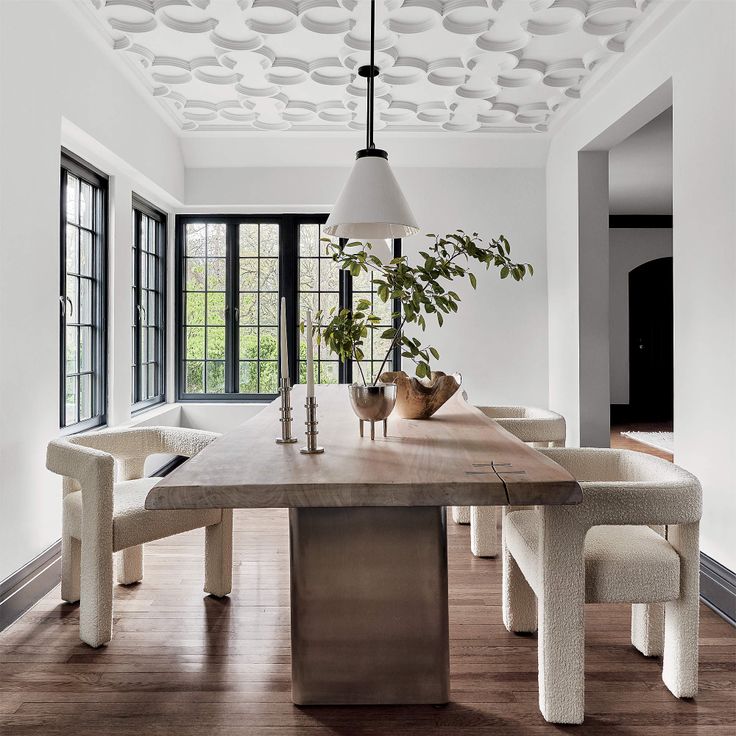 a dining room table with chairs and a potted plant on top of it in front of large windows