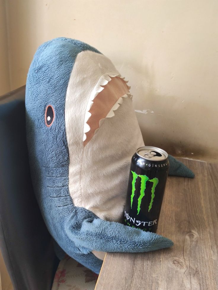 a blue stuffed shark with a can of monster energy on it's back sitting at a table