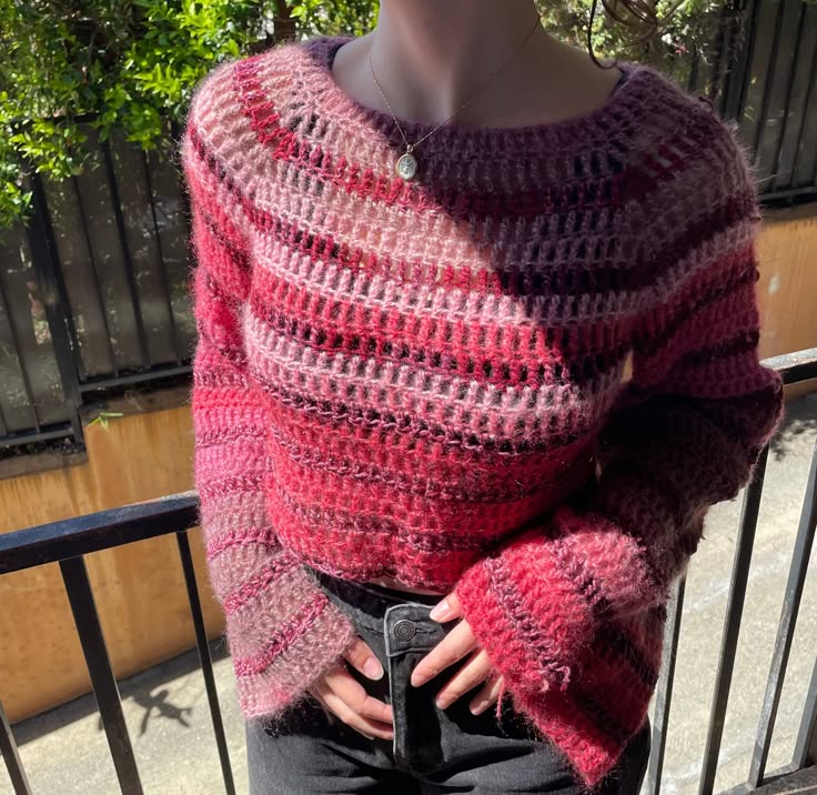 a woman wearing a pink and red sweater standing next to a metal fence with her hands in her pockets