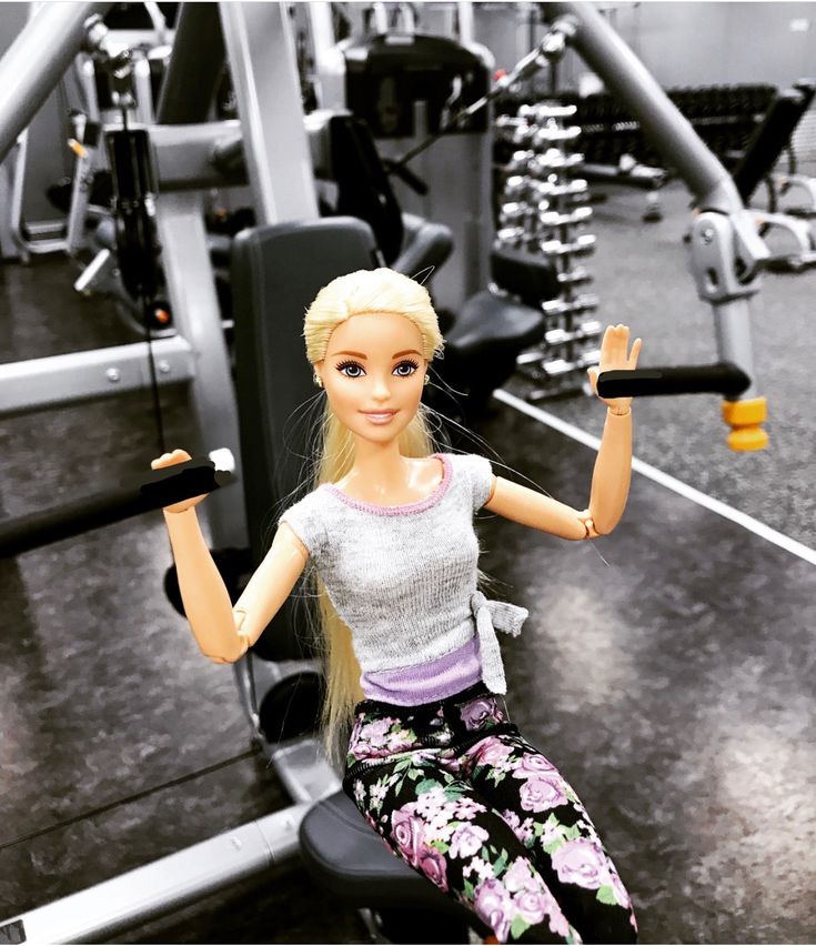 a barbie doll sitting on top of a bench holding a barbell in a gym