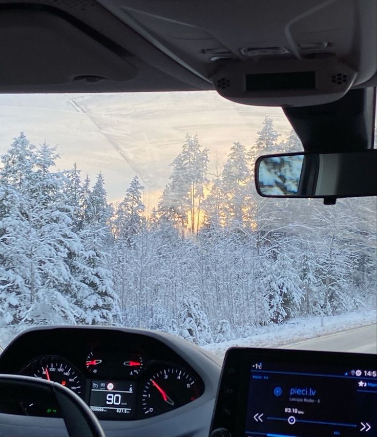 the dashboard of a car driving down a road in front of snow covered trees and evergreens