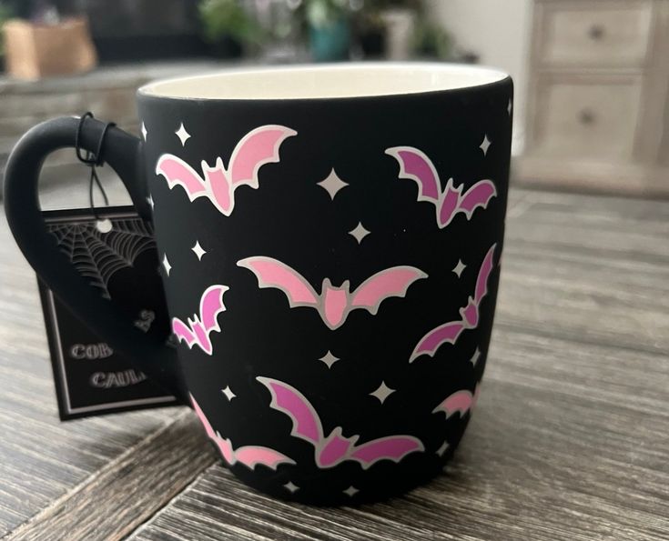 a black mug with pink bats and stars on it sitting on a wooden table next to a card