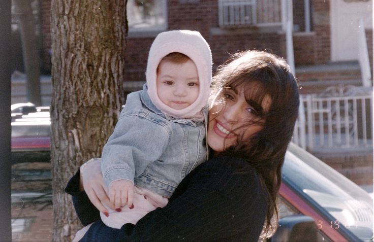 a woman holding a small child in her arms near a tree and parked car on the street