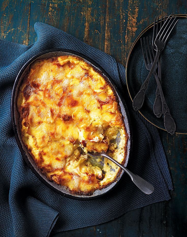 a casserole dish with cheese and vegetables on a blue cloth next to silverware