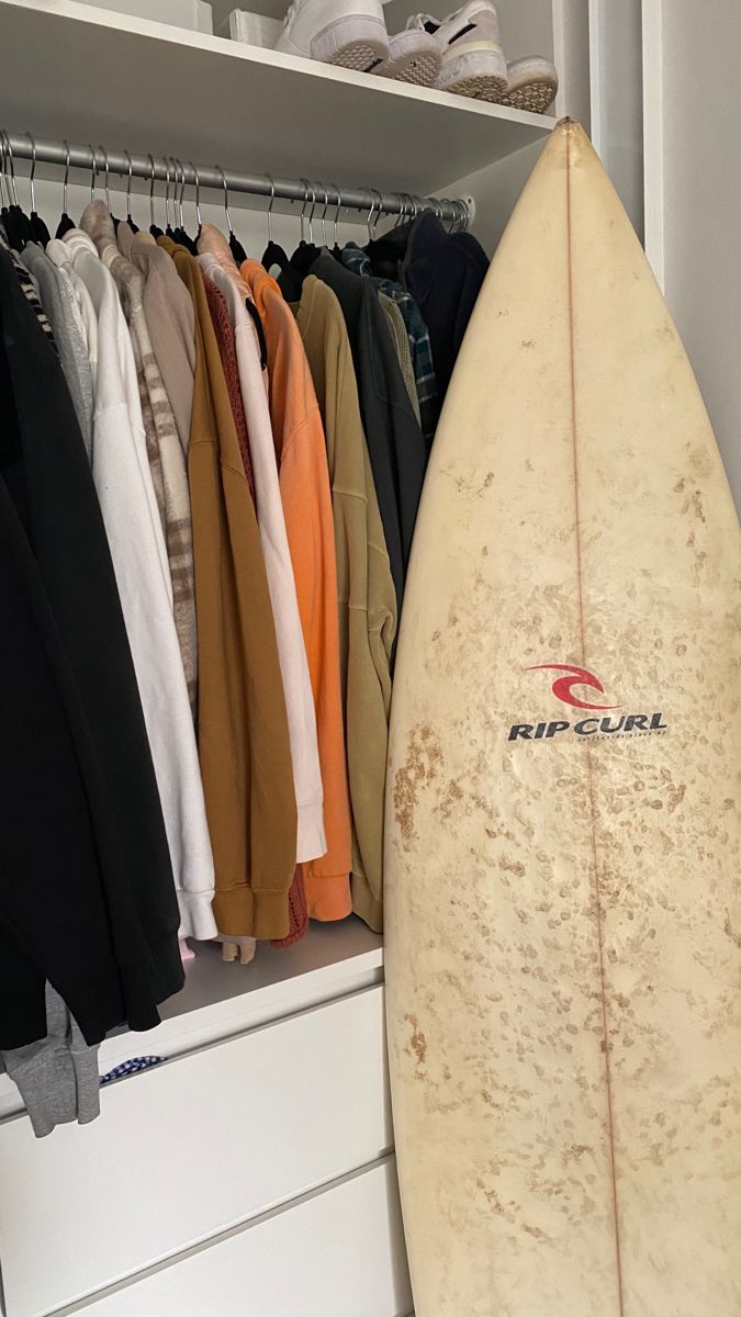 a surfboard sitting on top of a white shelf next to clothes hanging up in a closet