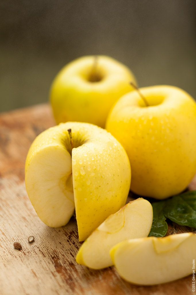 some yellow apples sitting on top of a wooden cutting board with leaves and water droplets