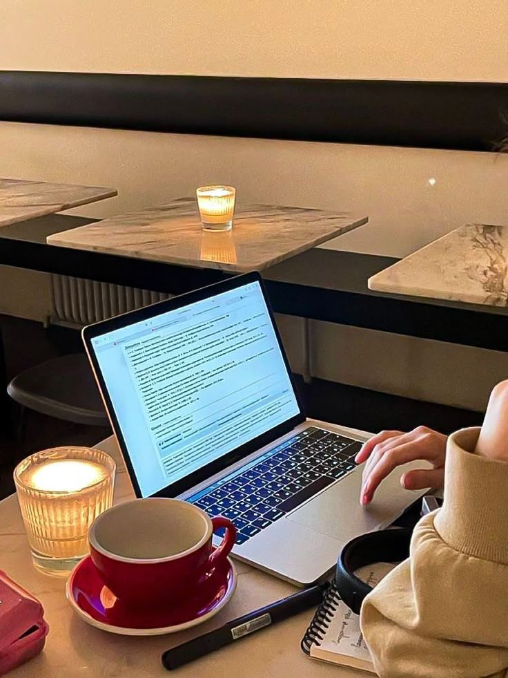 a woman sitting at a table with a laptop computer on her lap and coffee mug in front of her