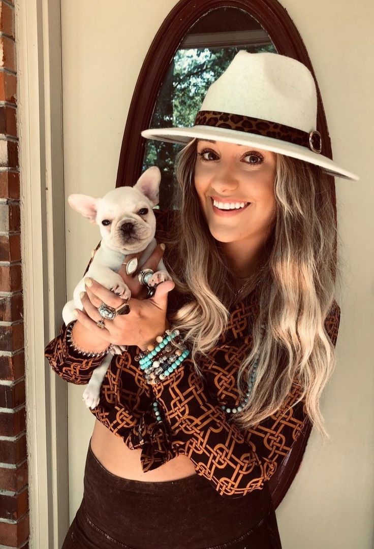 a woman holding a small dog in her arms while wearing a cowboy hat and sweater