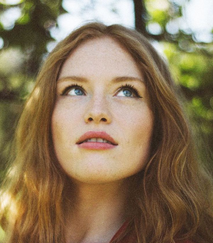 a woman with long red hair and blue eyes looks up into the sky while standing in front of trees