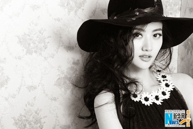 black and white photo of a woman wearing a hat with flowers on the collar, standing against a wall