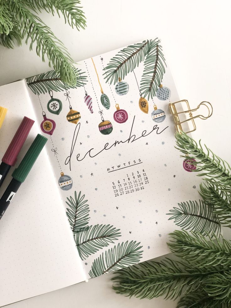 a calendar with christmas ornaments on it next to some pens and pencils in front of a pine tree