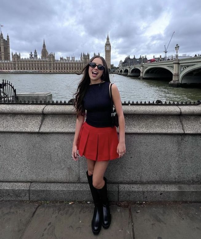 a woman in black shirt and red skirt standing by the water with big ben in the background