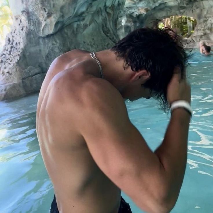 a shirtless man in the water with his hand on his head while standing next to a rock formation