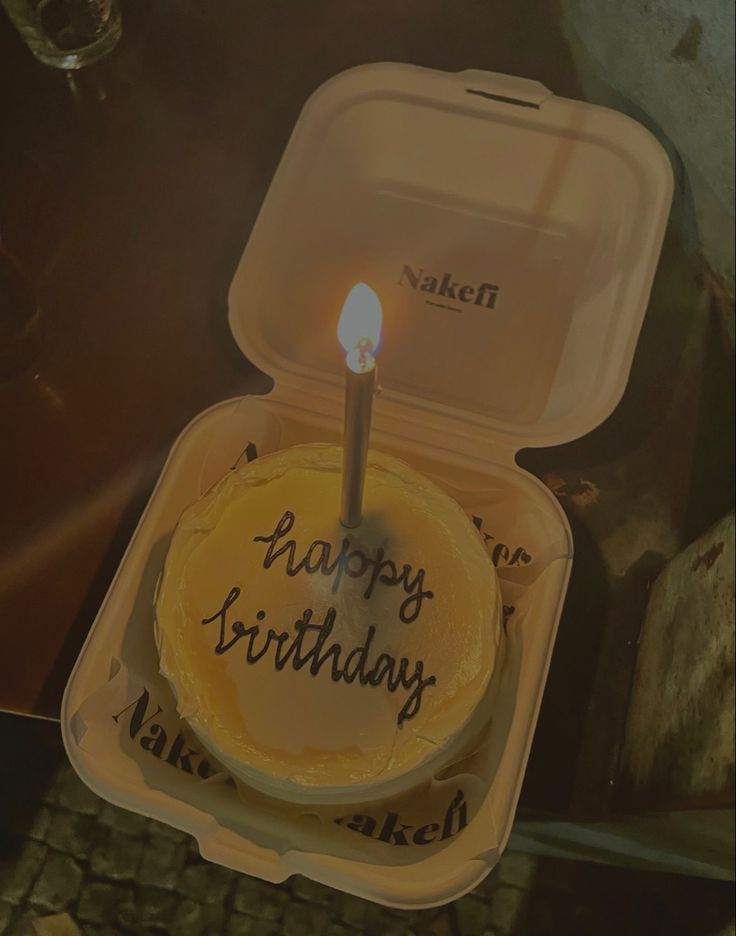 a birthday cake in a plastic container with a lit candle on it that says happy birthday