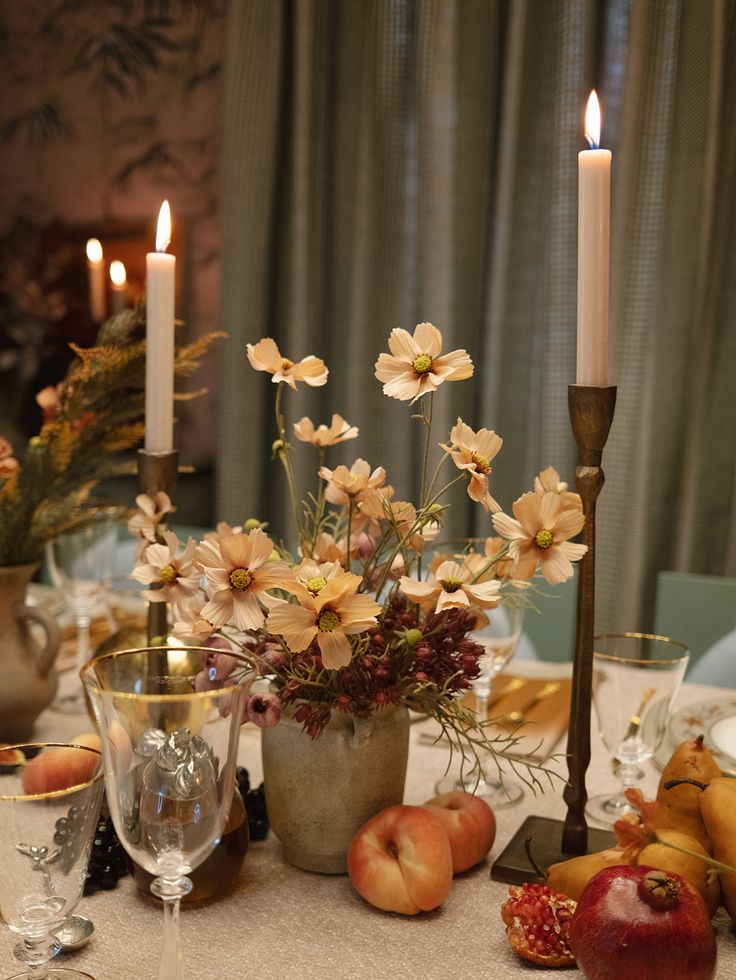 an arrangement of flowers and fruit on a table with two candles in the background,