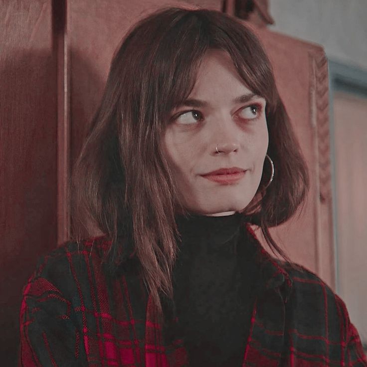 a woman with long hair wearing a red and black shirt standing in front of a wooden door