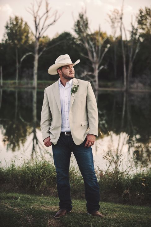 a man standing in front of a lake wearing a cowboy hat and suit with his hands in his pockets