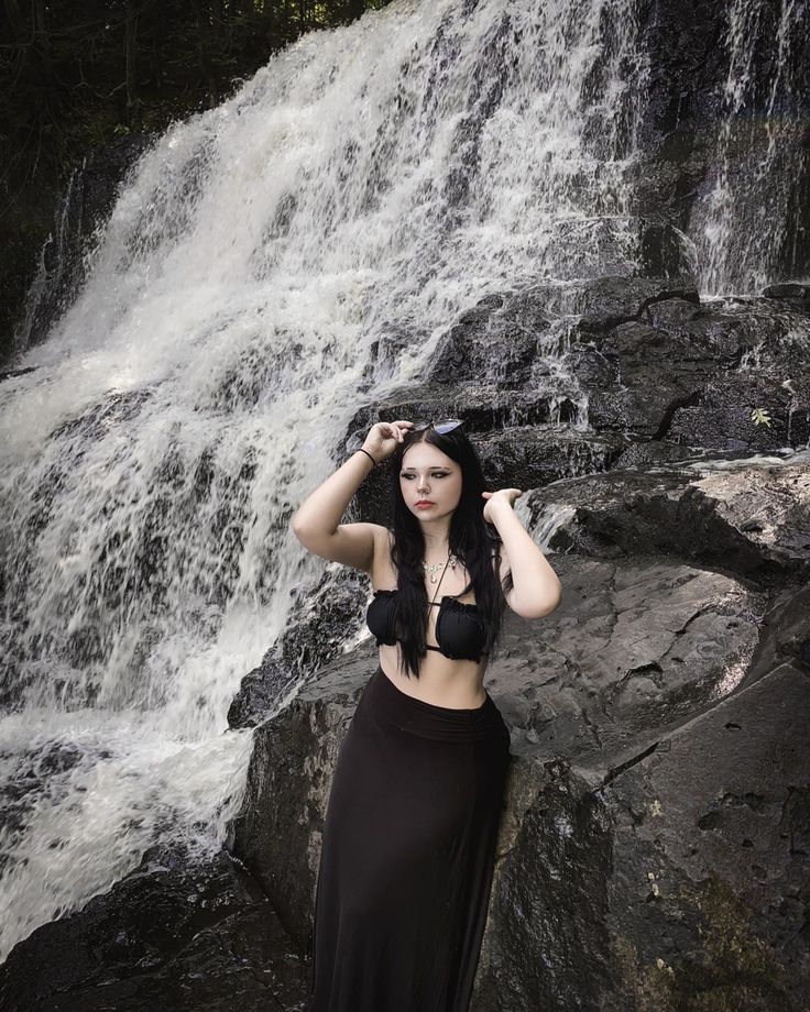 a woman standing in front of a waterfall with her hands on her head and wearing a black skirt