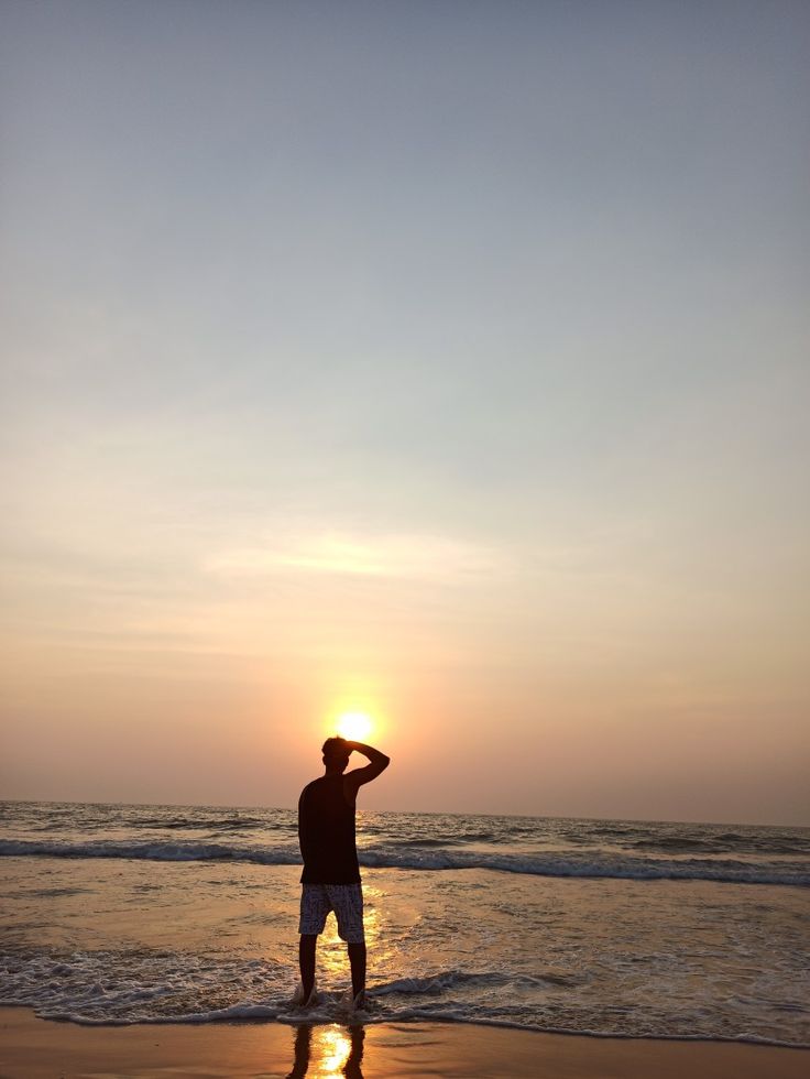 a man standing on top of a sandy beach next to the ocean under a sunset