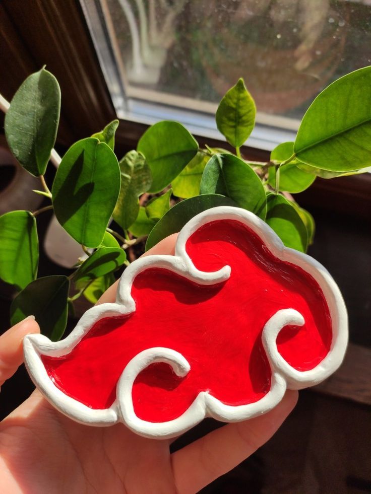 a hand holding a red and white ceramic object next to a green plant in front of a window