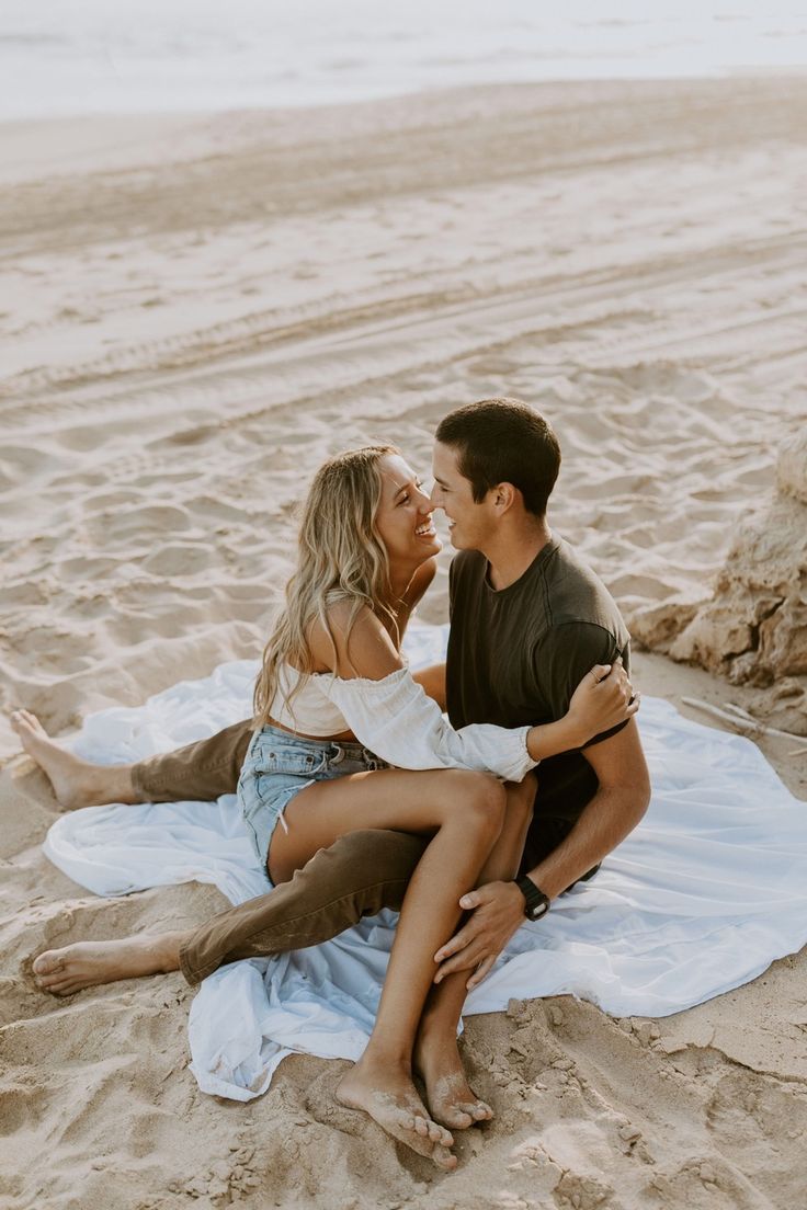 a man and woman sitting on top of a blanket in the sand at the beach