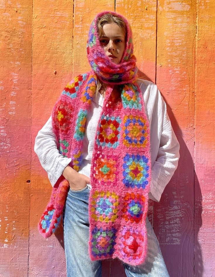 a woman wearing a pink crocheted scarf with flowers on it, standing in front of a wooden wall