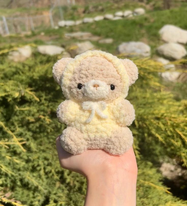 a small teddy bear sitting on top of someone's finger in front of some bushes