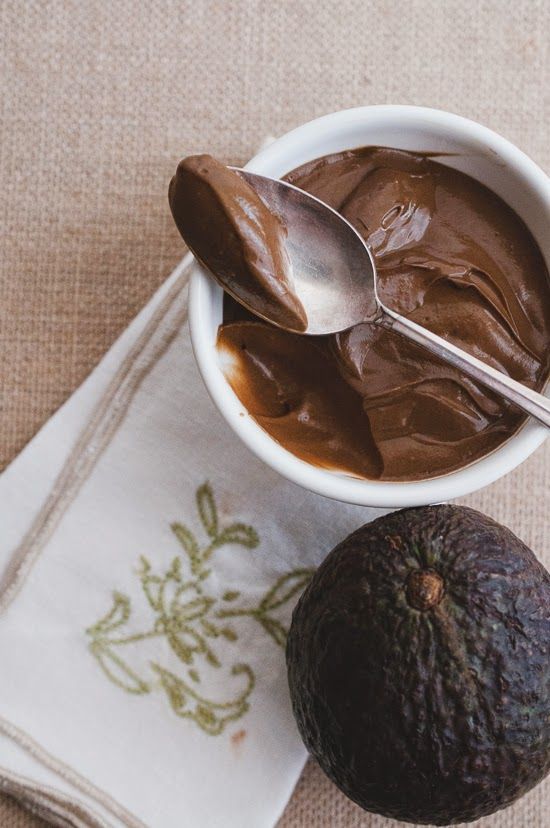 an avocado, spoon and chocolate spread in a white bowl on a napkin