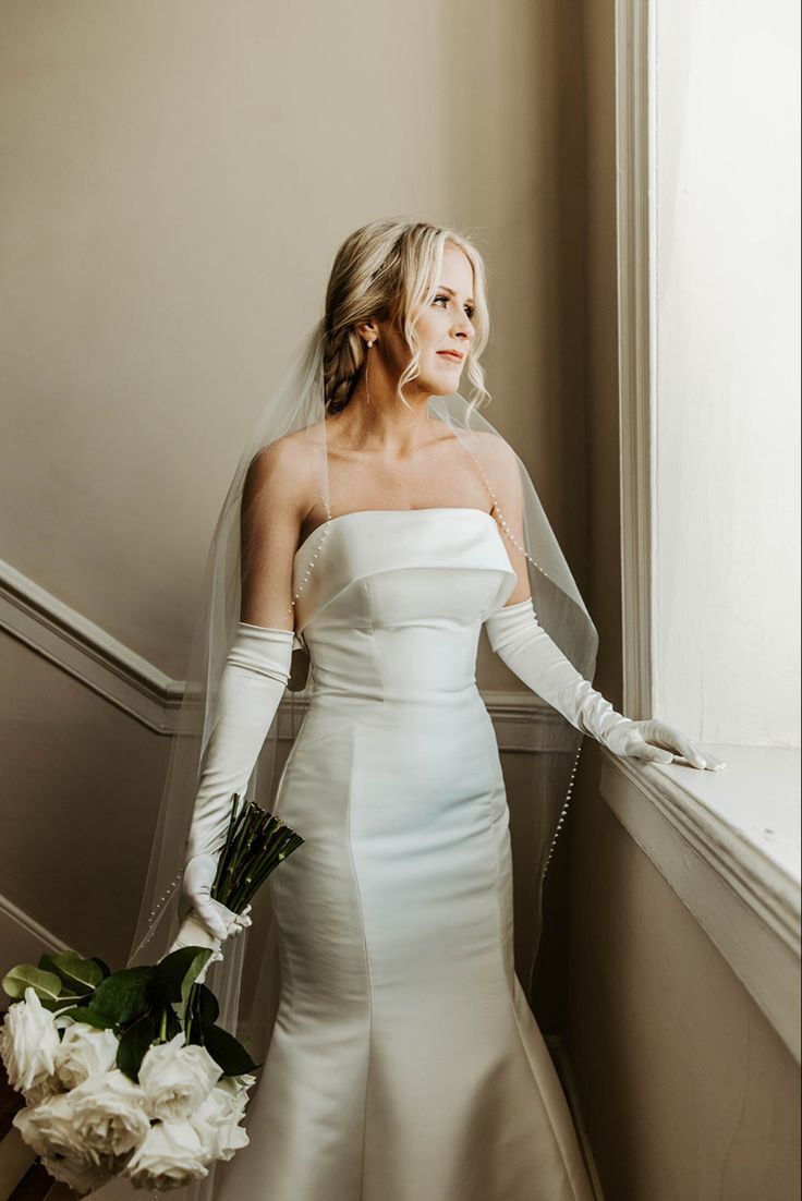 a woman in a wedding dress standing next to a window with her veil pulled back