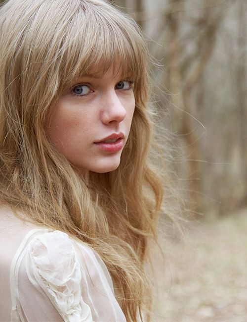 a woman with long blonde hair and blue eyes in the woods looking off into the distance