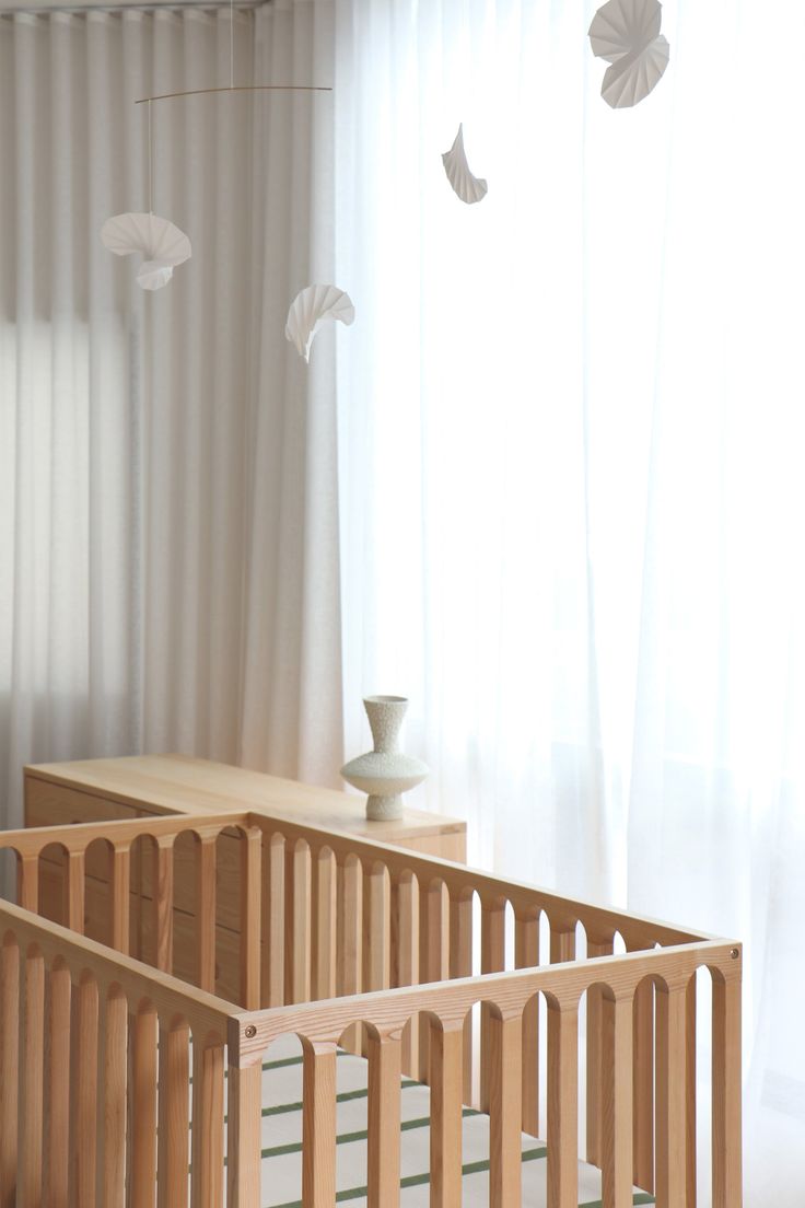 a baby crib in front of a window with white curtains and paper birds hanging from it