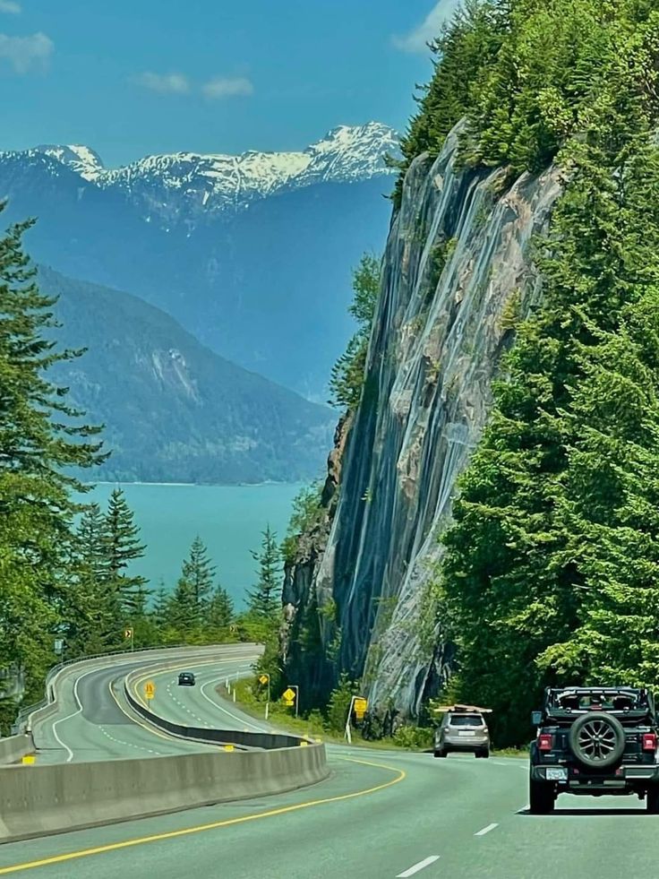 two cars driving down the road next to some mountains and trees with snow capped peaks in the distance