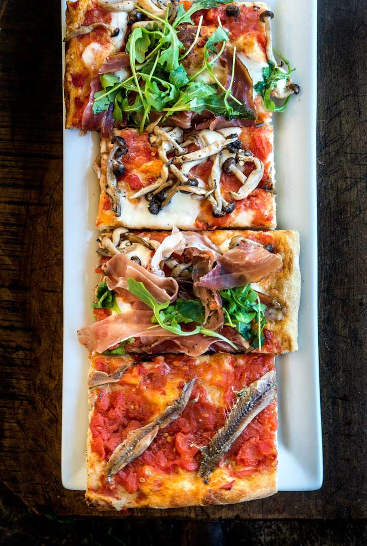 three slices of pizza on a white plate with greens and other toppings sitting on a wooden table