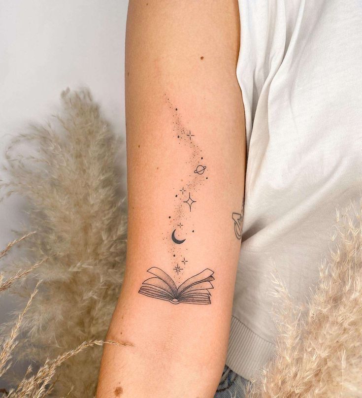 a woman's arm with a book and stars tattoo on her left arm, next to some dry grass