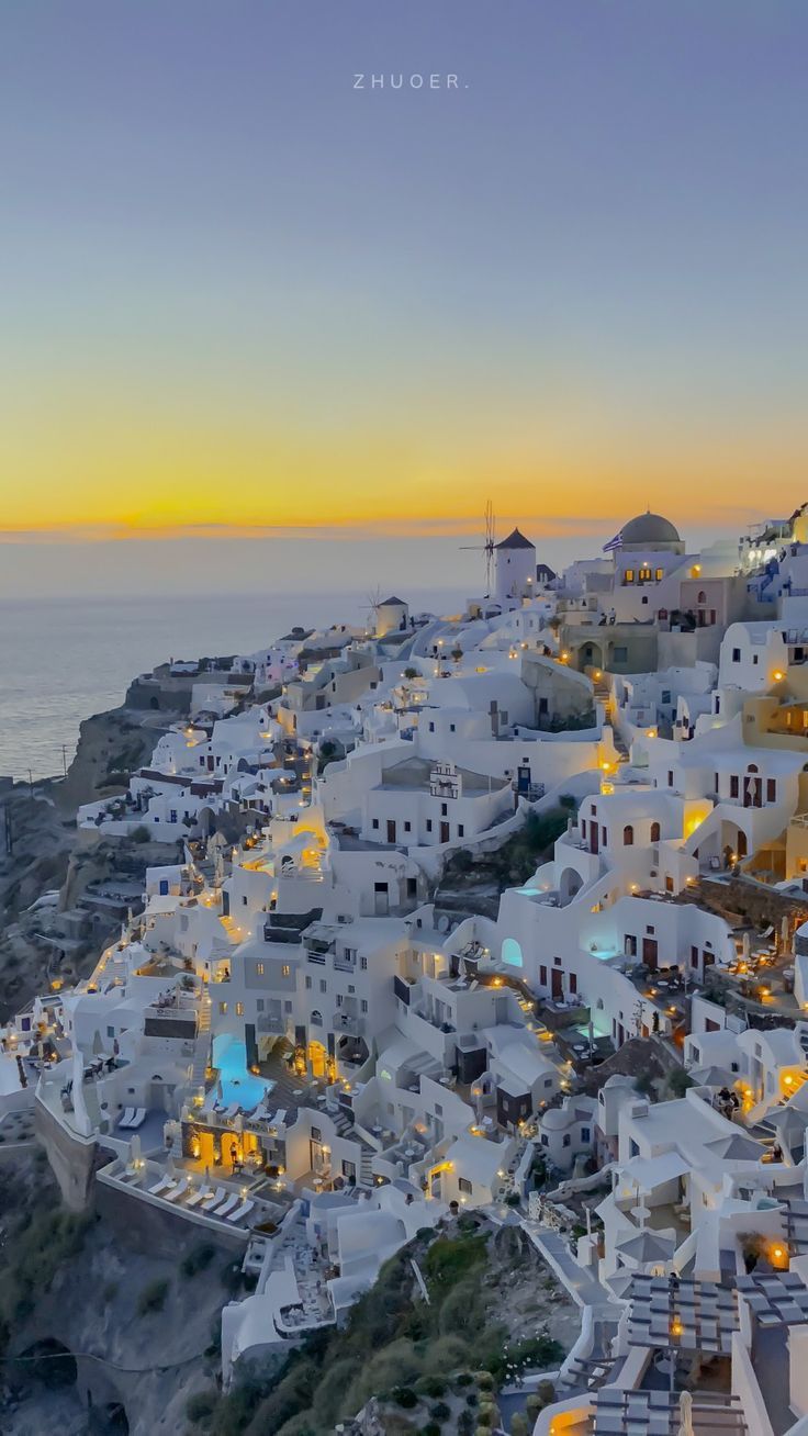 the white buildings are lit up at night on the cliff above the water and in front of the ocean