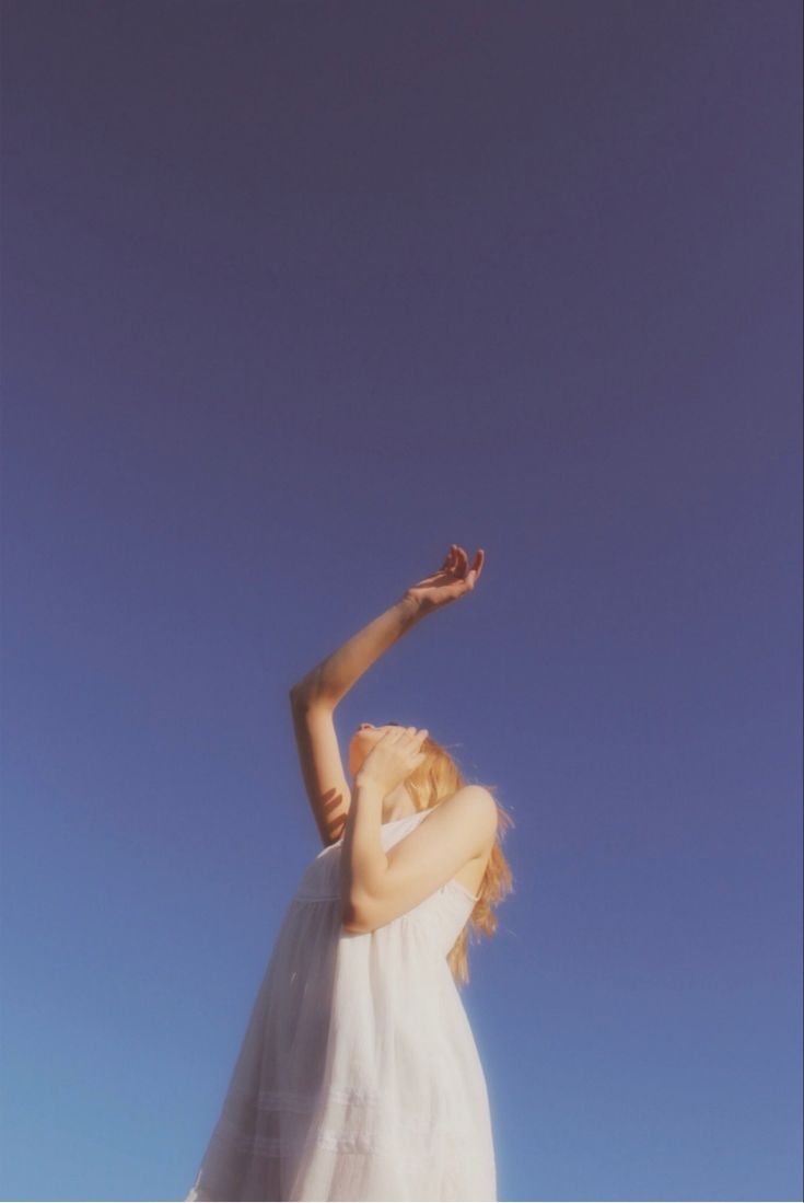 a woman in a white dress flying a kite high up in the air with her arms outstretched