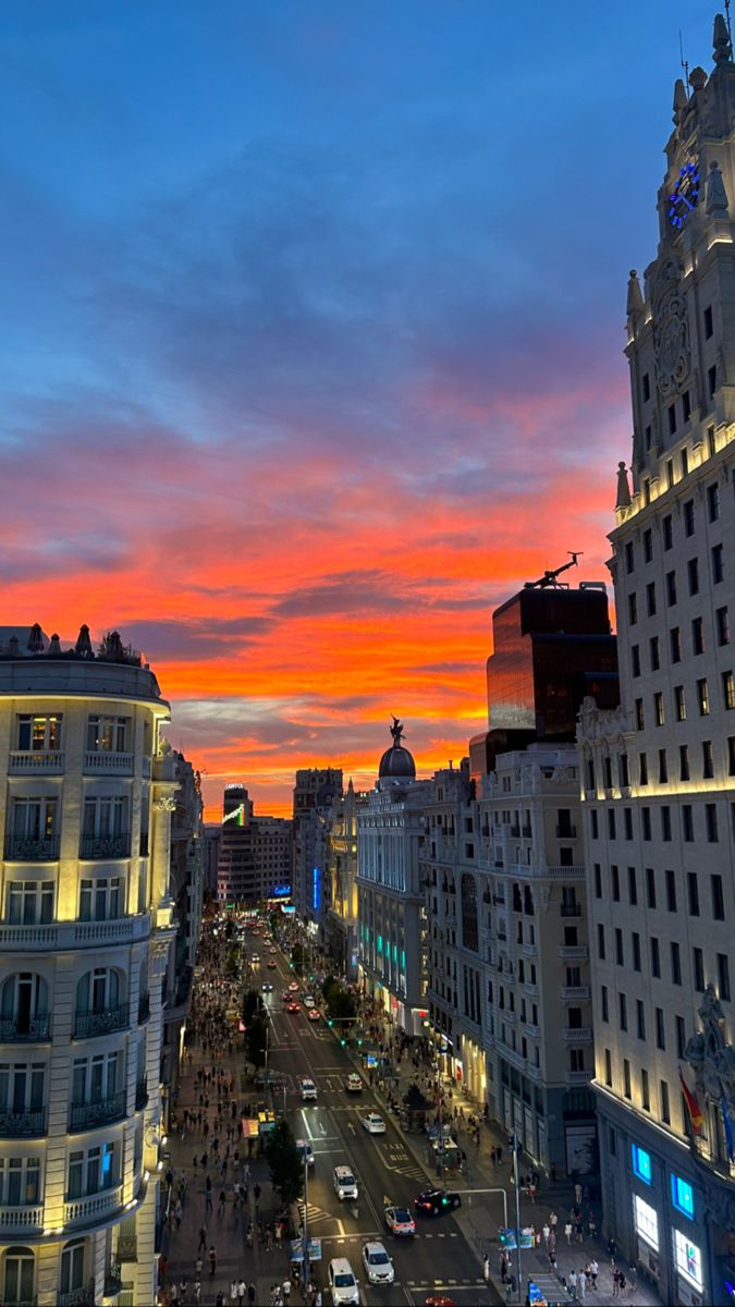 the sun is setting over a city street with tall buildings and cars driving down it