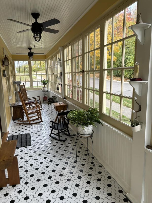 an empty porch with rocking chairs and potted plants on the floor, in front of large windows