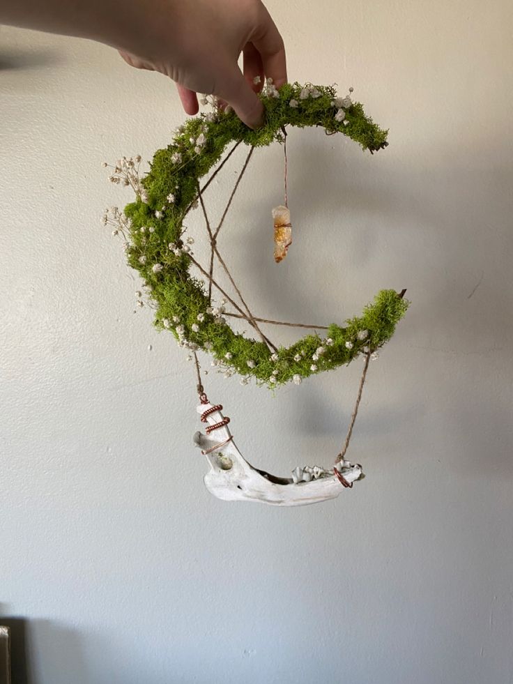 a hand holding a piece of art made out of moss and beads with the shape of a wreath hanging from it's side