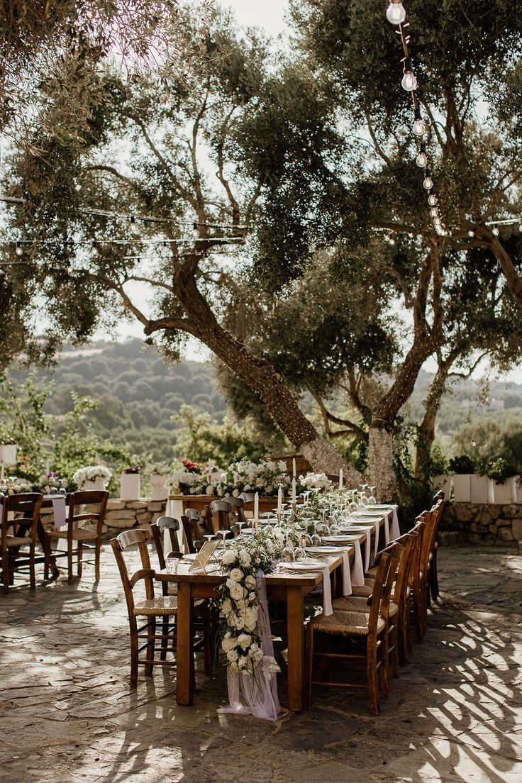 an outdoor dining table set up with flowers and greenery on the tables, surrounded by trees