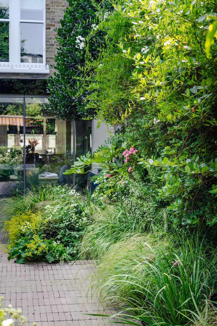 a garden with lots of green plants and flowers on the side of a brick walkway