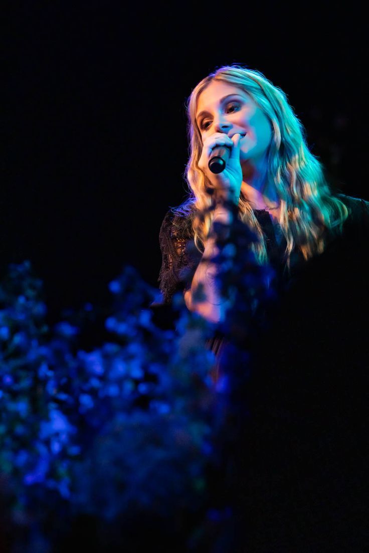 a woman singing into a microphone in front of blue and green lights on a black background